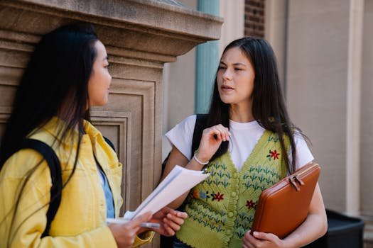 Two female students conversing on a college campus, sharing documents and ideas.